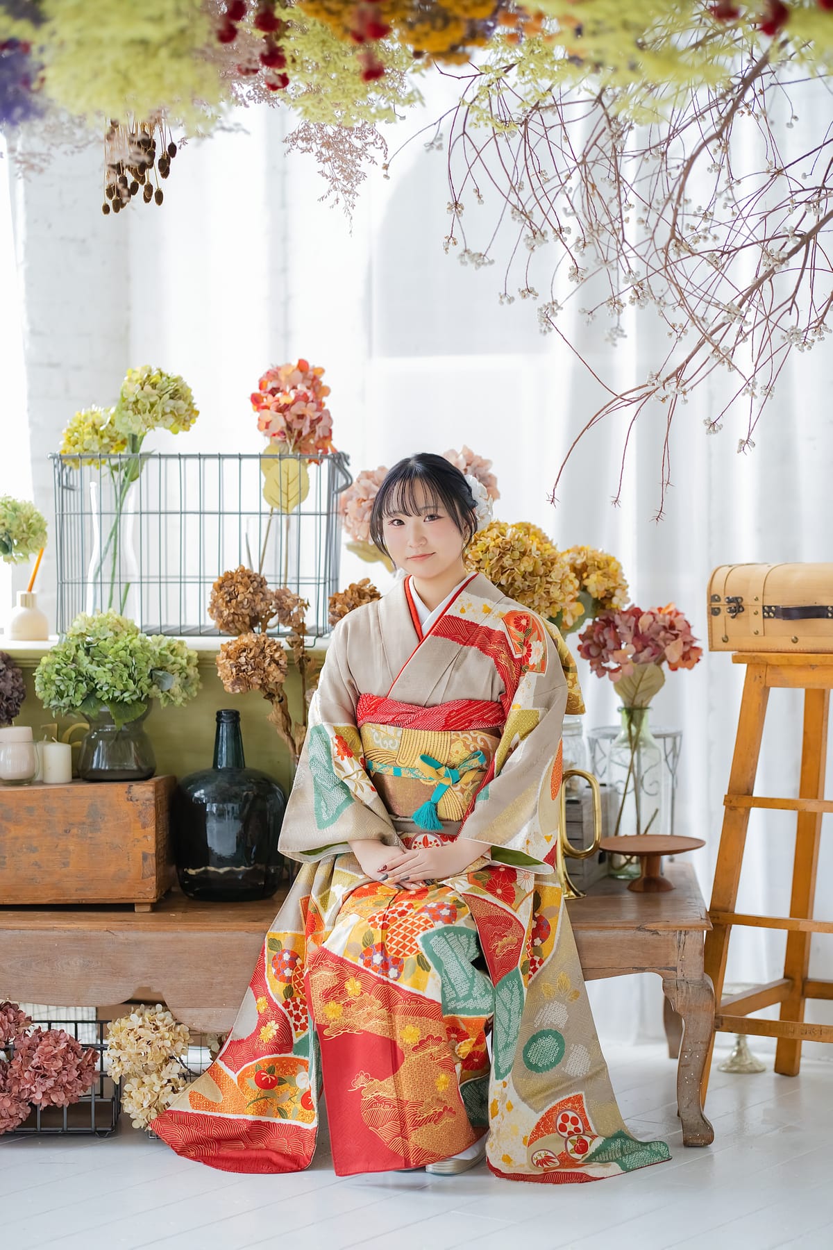 生成りに朱の花の振袖。花を並べた空間で座った姿