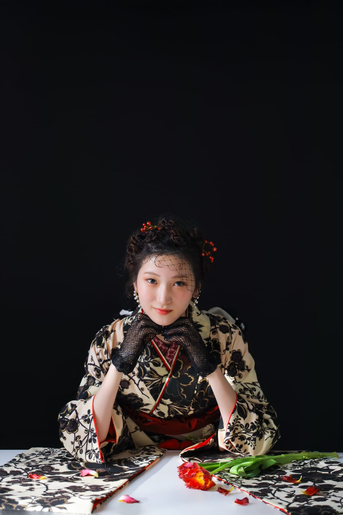 生成りに黒い花の振袖。暗い背景で机に手を置いた上半身
