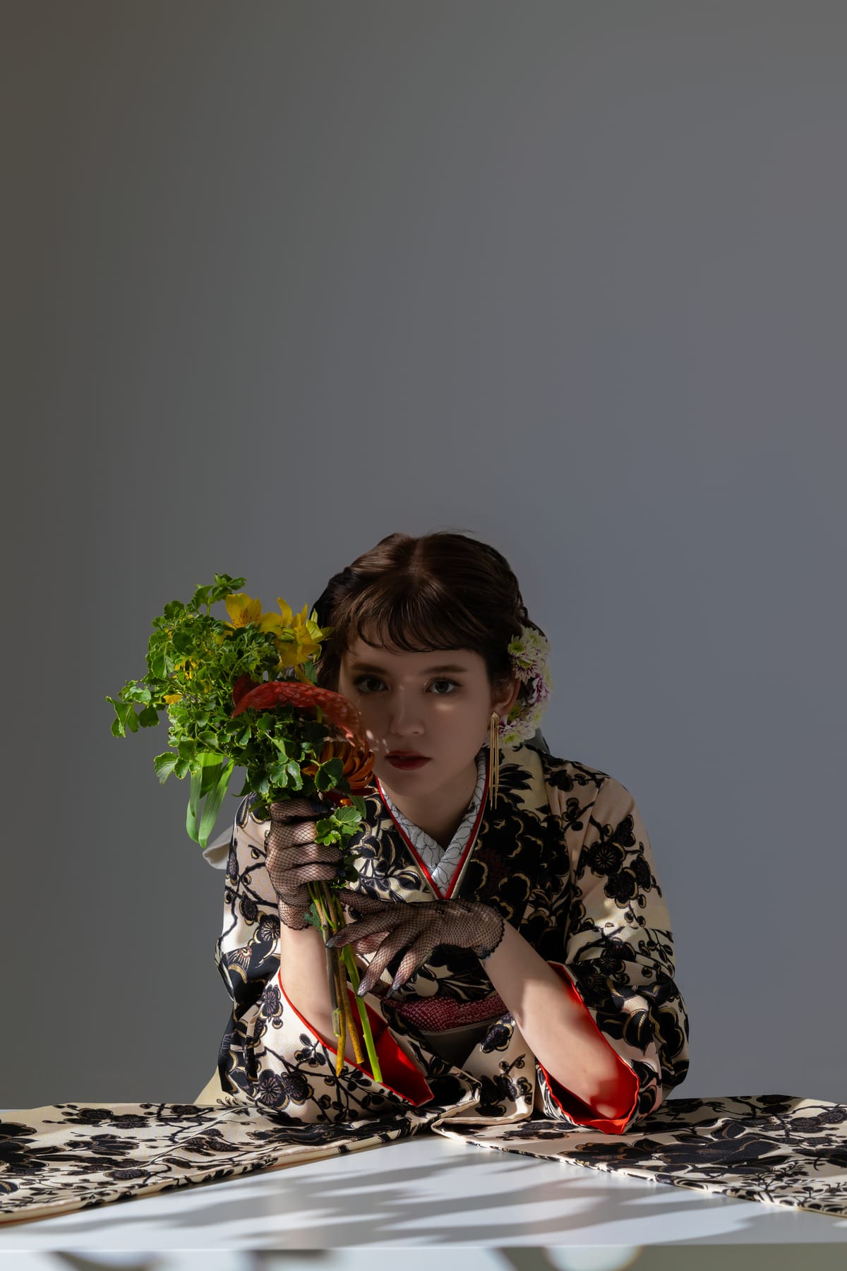 生成りに黒い花の振袖。花束を持って机に肘をついた上半身