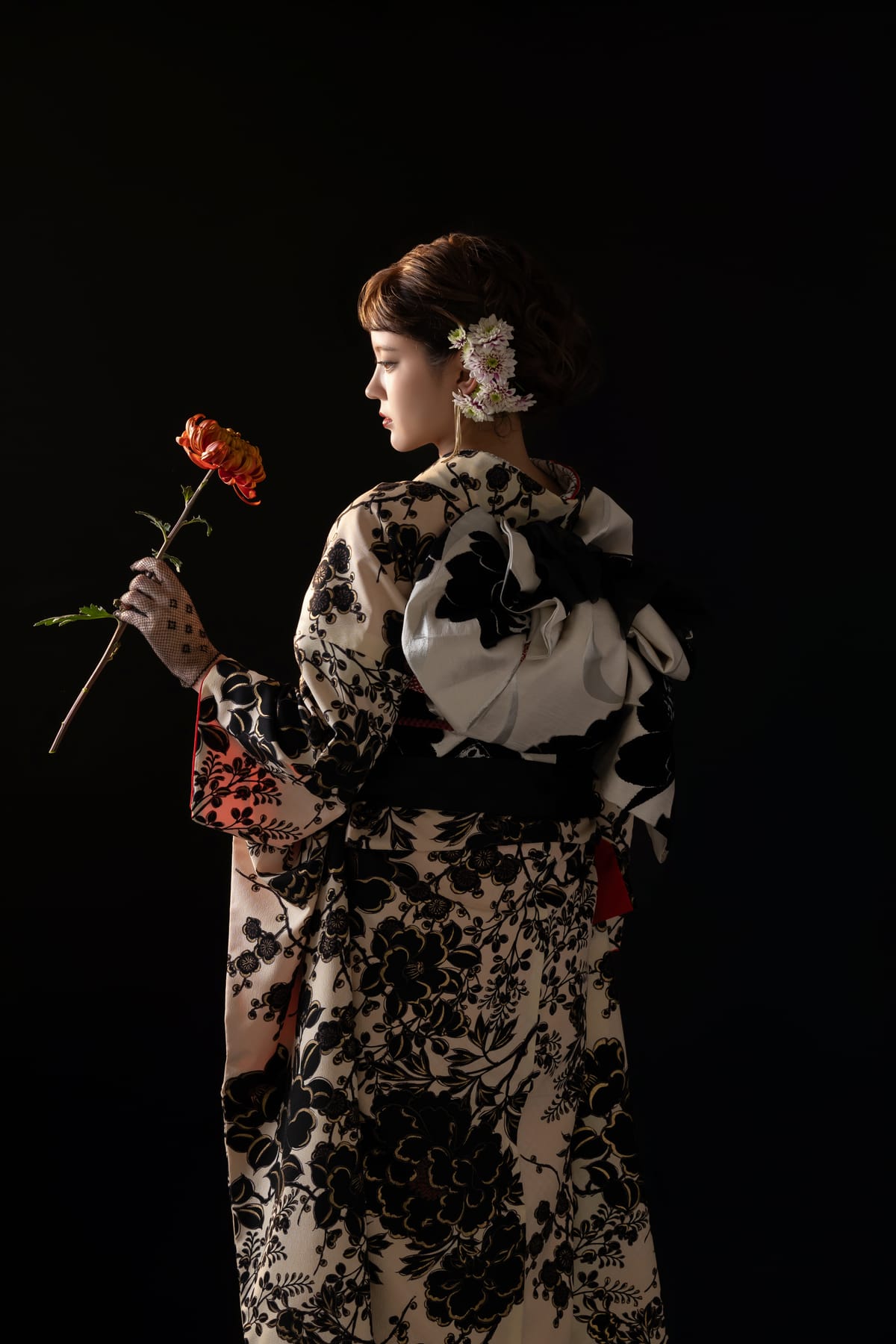 黒い背景紙の前で、生成りに黒い花の振袖を着て花を手にした後ろ姿