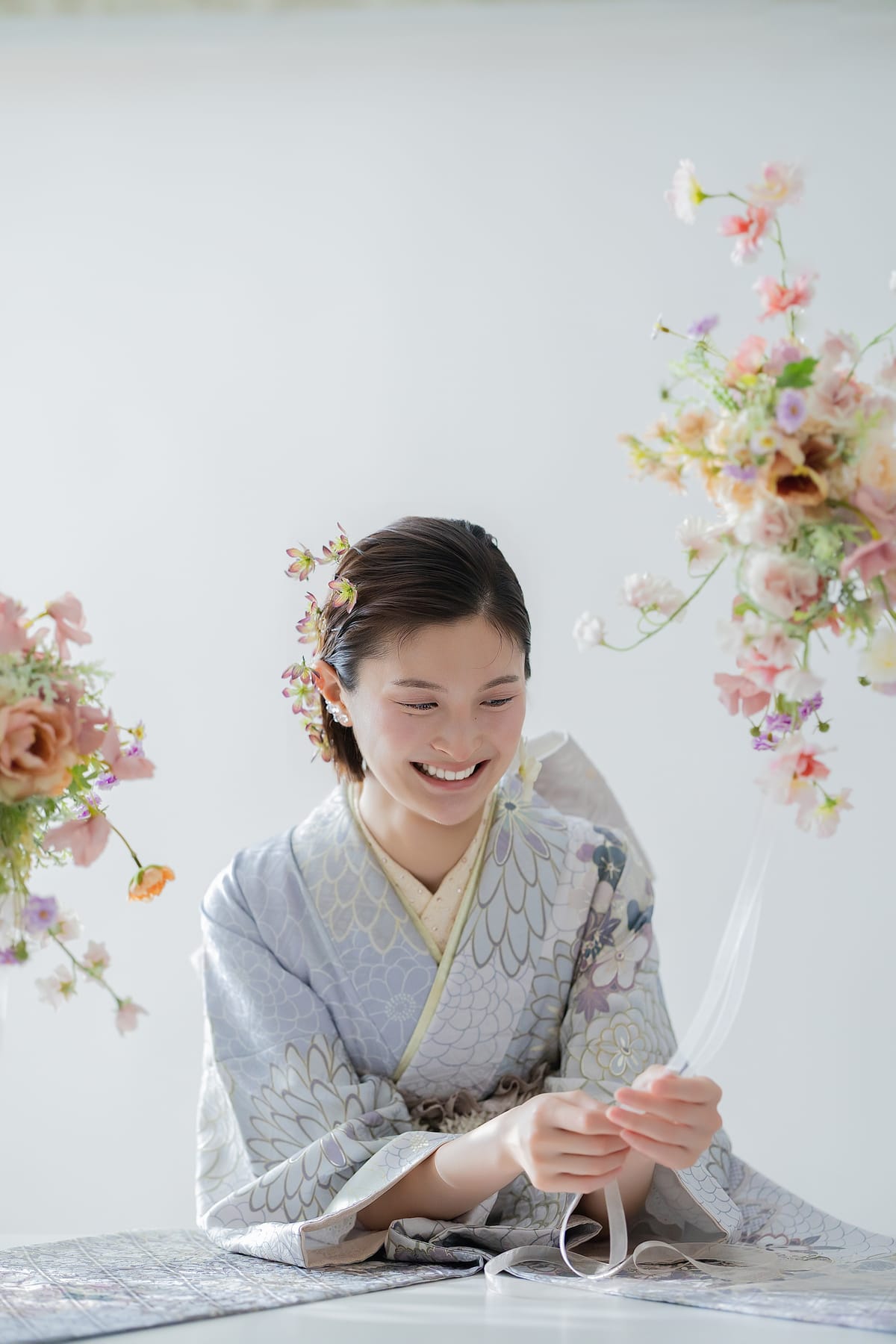白い空間で花に囲まれ、まとめ髪の横顔が見える座り姿